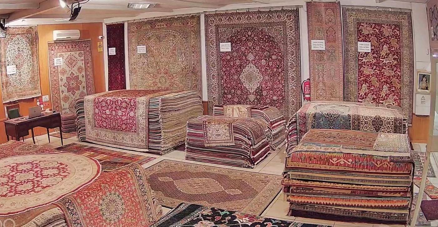 Sala de exposição da Galeria Imperial, com tapetes de seda pendurados na parede e pilhas de tapetes no chão